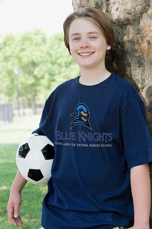 A young man wearing a Blue Knights t-shirt and carrying a soccer ball poses against a tree.